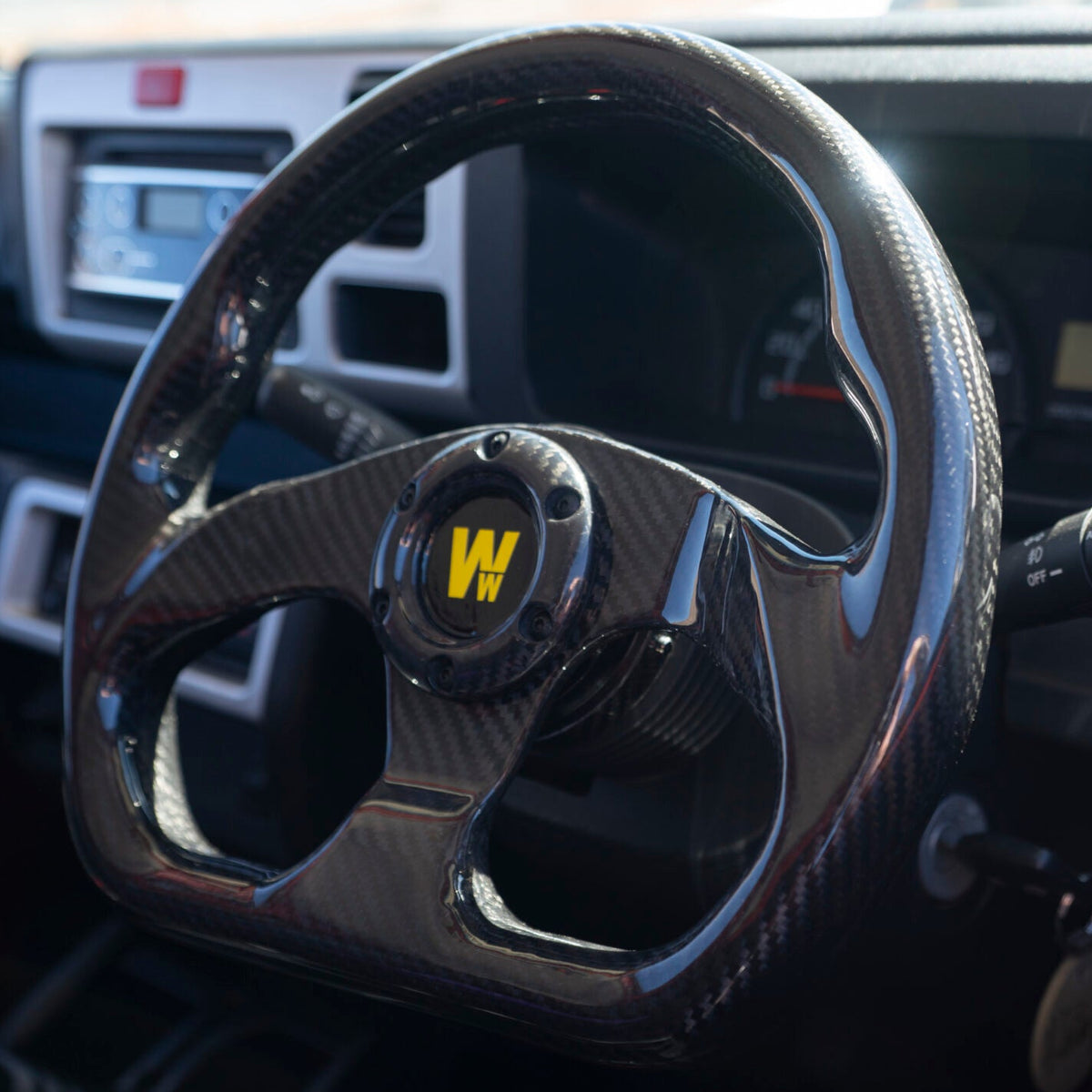 A close-up of a black steering wheel with a black center and a yellow 'W' logo, installed inside a vehicle cabin with a blurred outdoor background visible through the windshield.